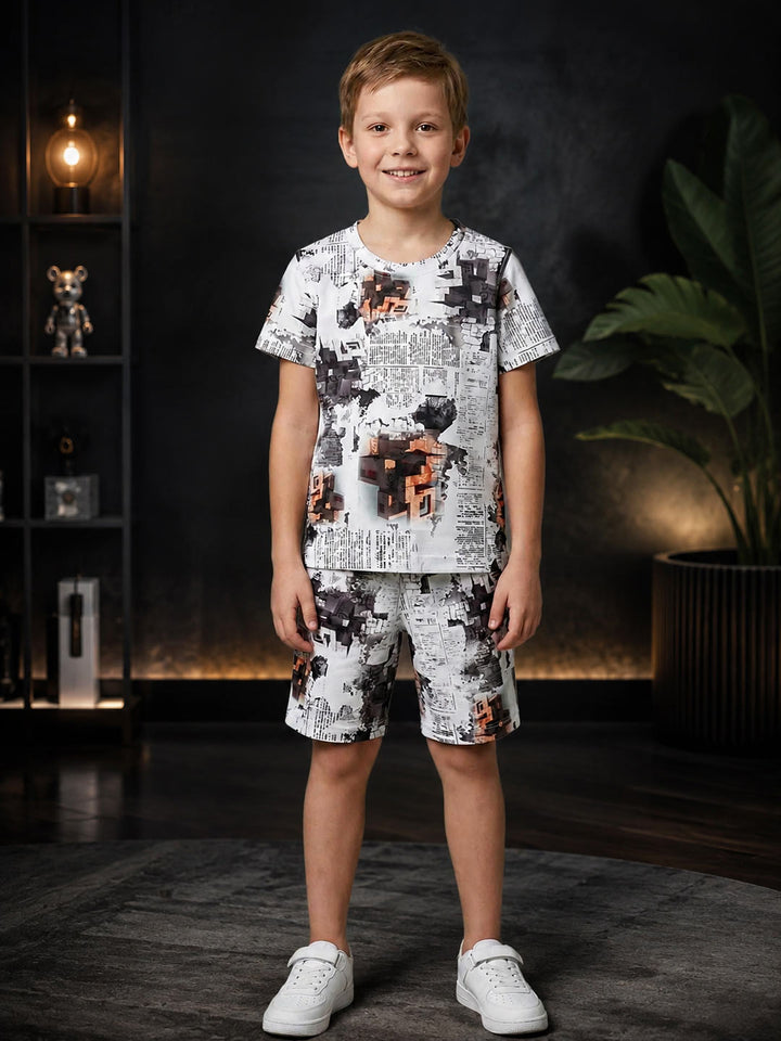 A young boy stands in a dark, modern room wearing a matching two-piece pajama set with a white background and black, gray, and orange abstract pixel-art style graphics. The set includes a short-sleeve t-shirt and shorts, paired with white sneakers.