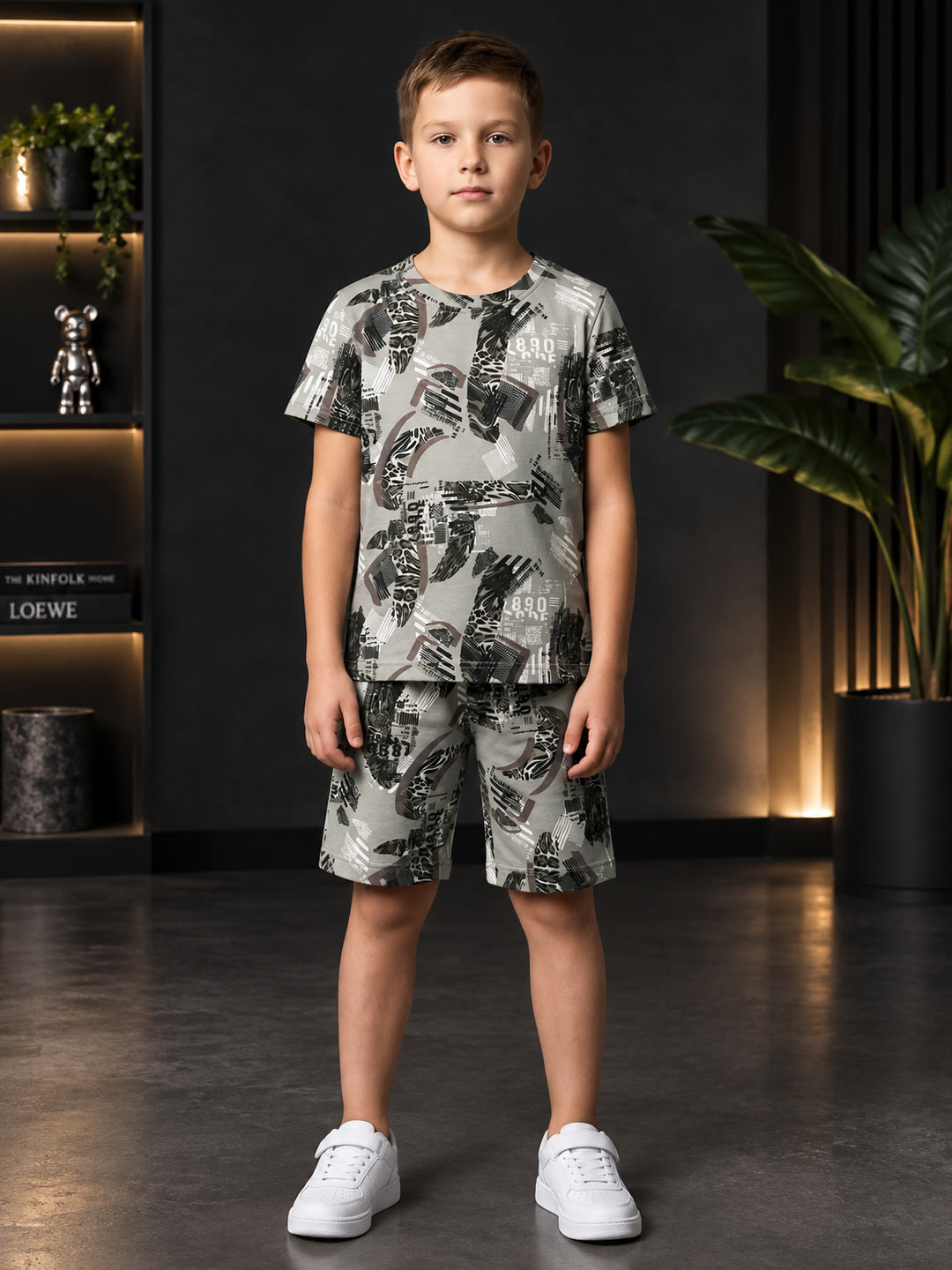 A young boy stands in a modern room wearing a matching short-sleeve t-shirt and shorts set with a black and gray palm leaf pattern on a light gray background, paired with white sneakers.