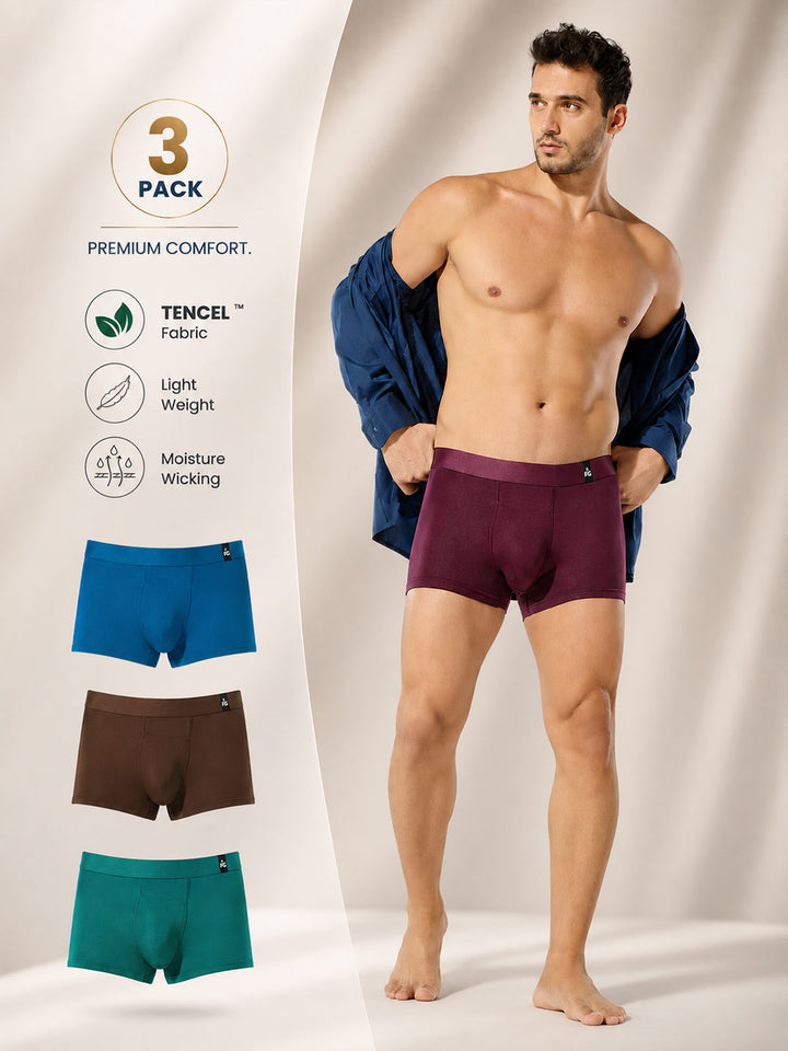 A male model stands in a studio wearing a pair of plum boxer briefs, with his navy jacket off his shoulders. On the left, a vertical panel shows a '3 PACK' badge, product features with TENCEL fabric icons, and three additional color options: blue, brown, and teal.