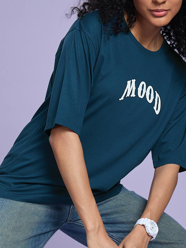 Person wearing a teal t-shirt with 'MOOD' printed on it against a light purple background