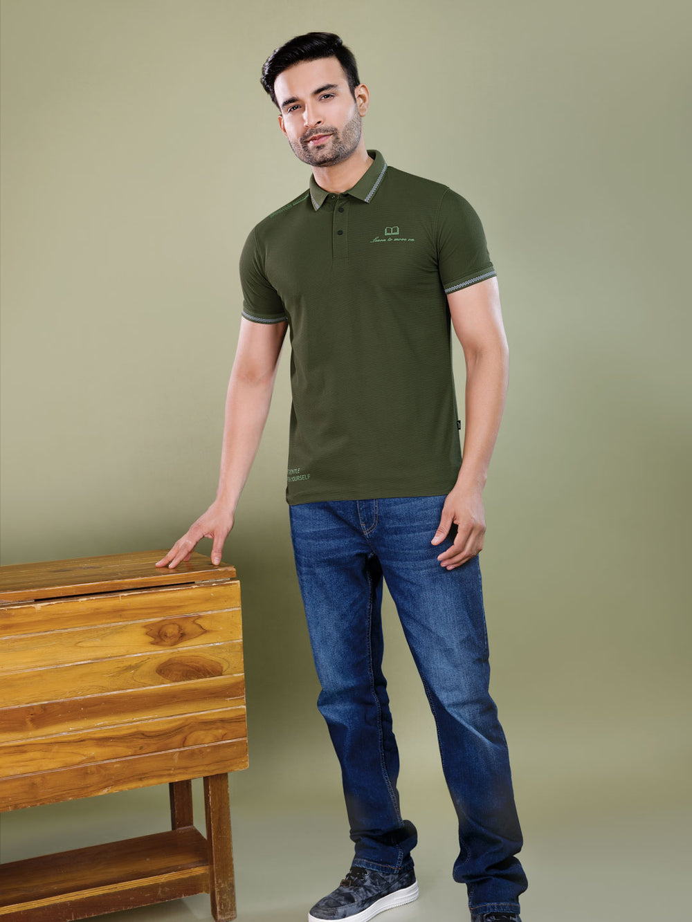 Man wearing a green polo shirt and blue jeans standing next to a wooden bench against a plain background