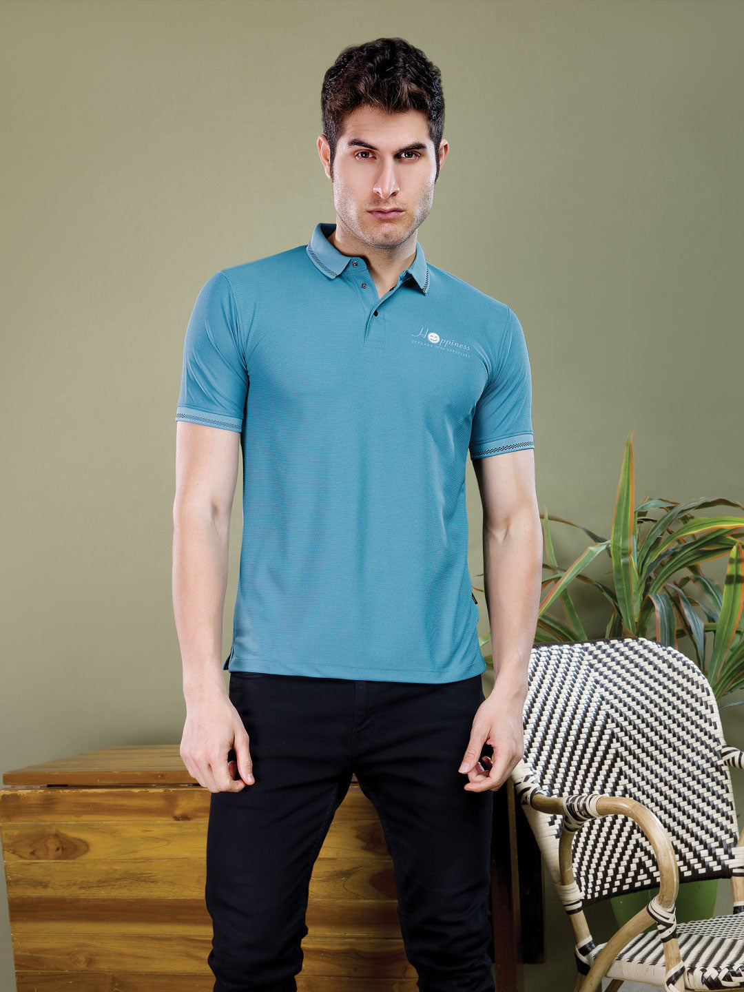 Man wearing a blue polo shirt standing in a room with a chair and plant.