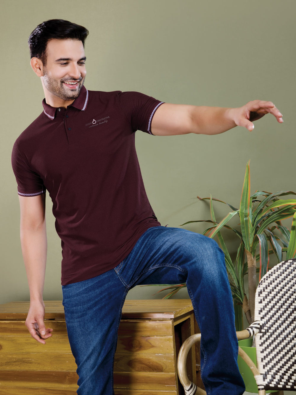 Man wearing a maroon polo shirt with a logo, sitting on a wooden stool indoors.