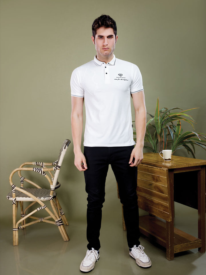 Man wearing a white polo shirt with a logo in a room with a wooden desk and chair.