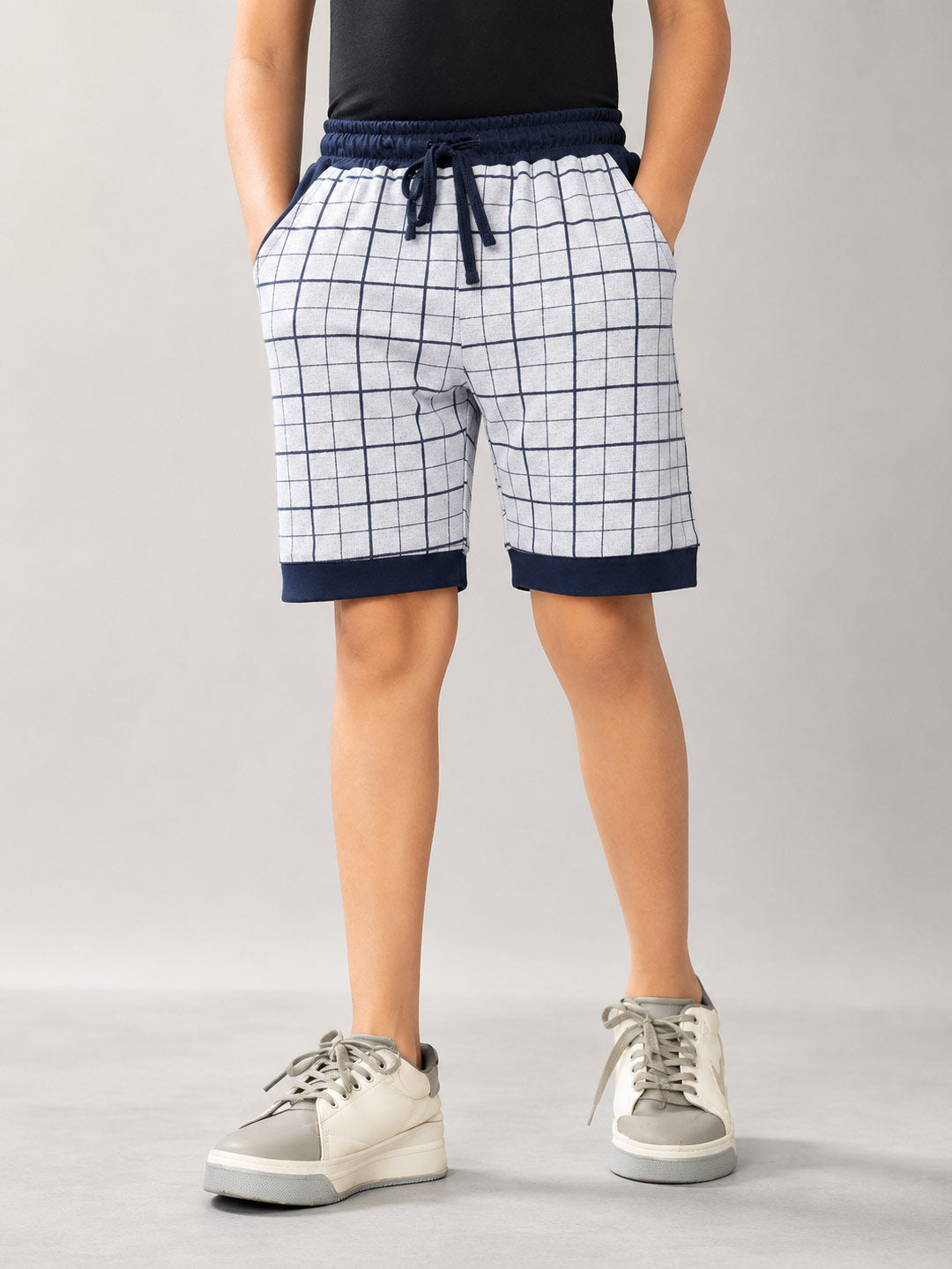 A person wearing white shorts with a navy blue grid pattern, a navy blue drawstring waist, and navy blue ribbed hems, paired with a black top and white sneakers, shown from the waist down on a light gray studio background.fitzone-105 shorts for boys color_dark-navy-blue