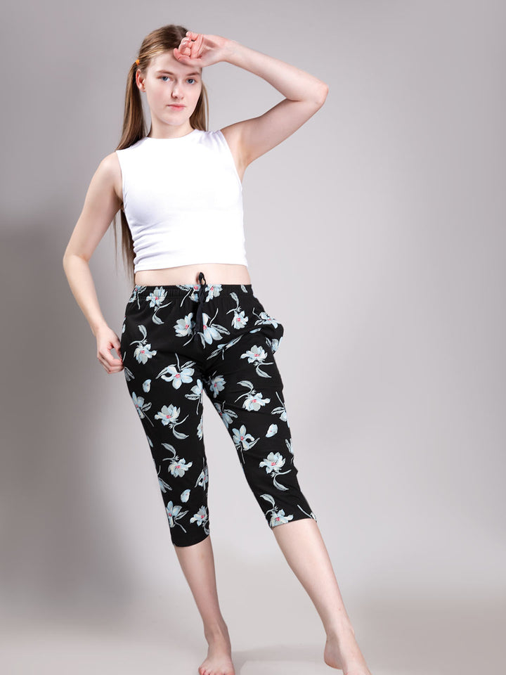 A woman modeling black capri pants with a white floral print, paired with a white sleeveless crop top, against a plain gray studio background.