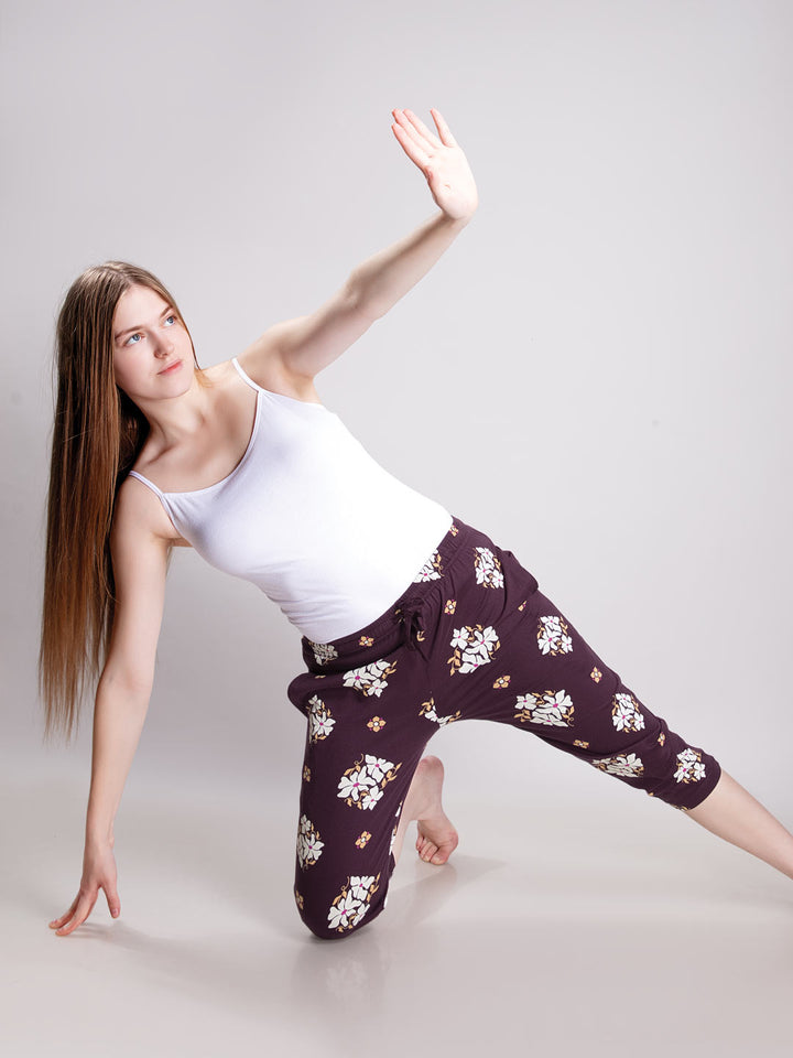 A woman in a white tank top and dark purple floral capri pants poses dynamically in a side stretch on a light gray studio background.
