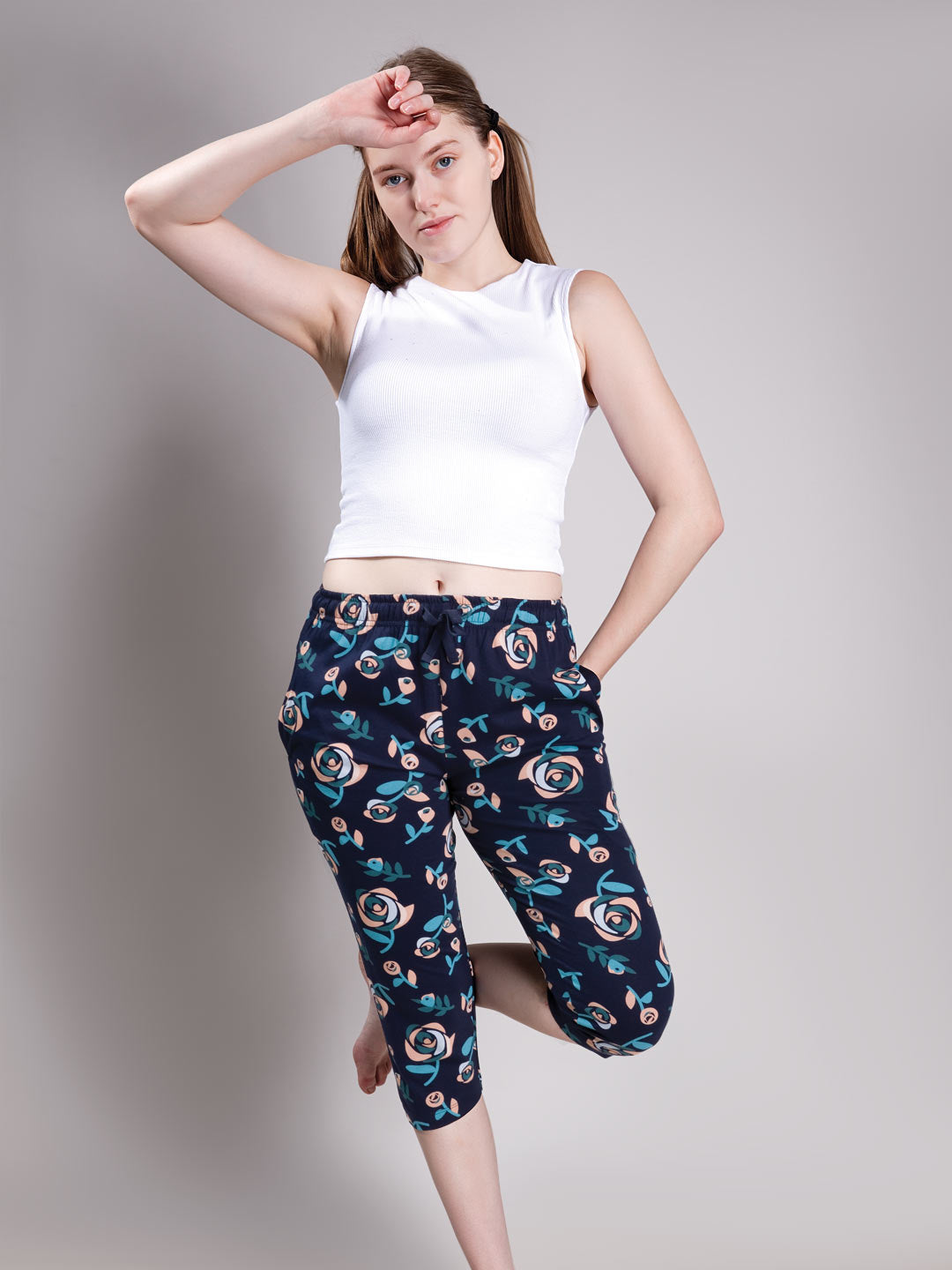 A woman modeling a pair of navy blue capri pants with a colorful floral pattern, paired with a white sleeveless crop top, against a plain gray studio background.