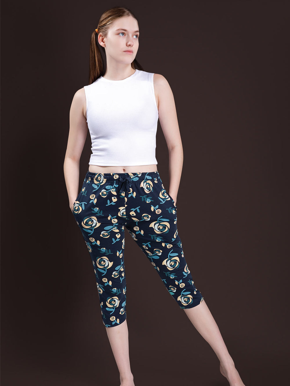 A woman modeling a white sleeveless crop top and dark navy capri pants with a teal and cream floral pattern, posed against a solid dark brown studio background.