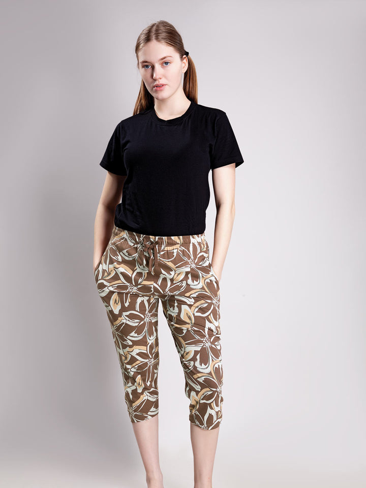 A woman modeling a black short-sleeve t-shirt and brown cropped pants with a beige and white botanical leaf pattern, standing against a light gray studio background.