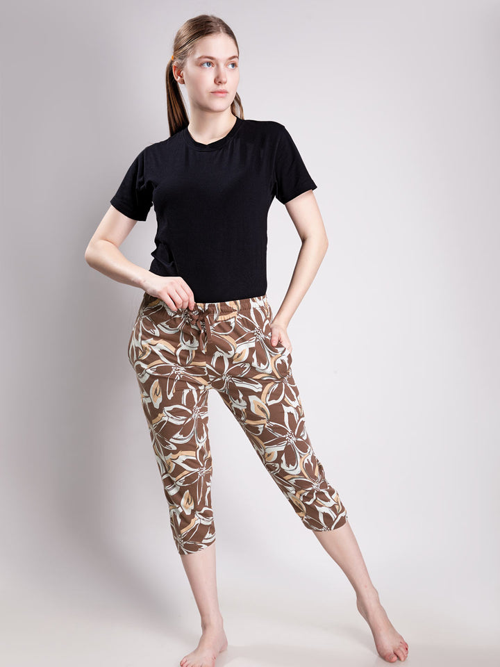 A woman modeling a pair of brown and beige floral print capri pants with a drawstring waist, paired with a black t-shirt, against a light gray studio background.