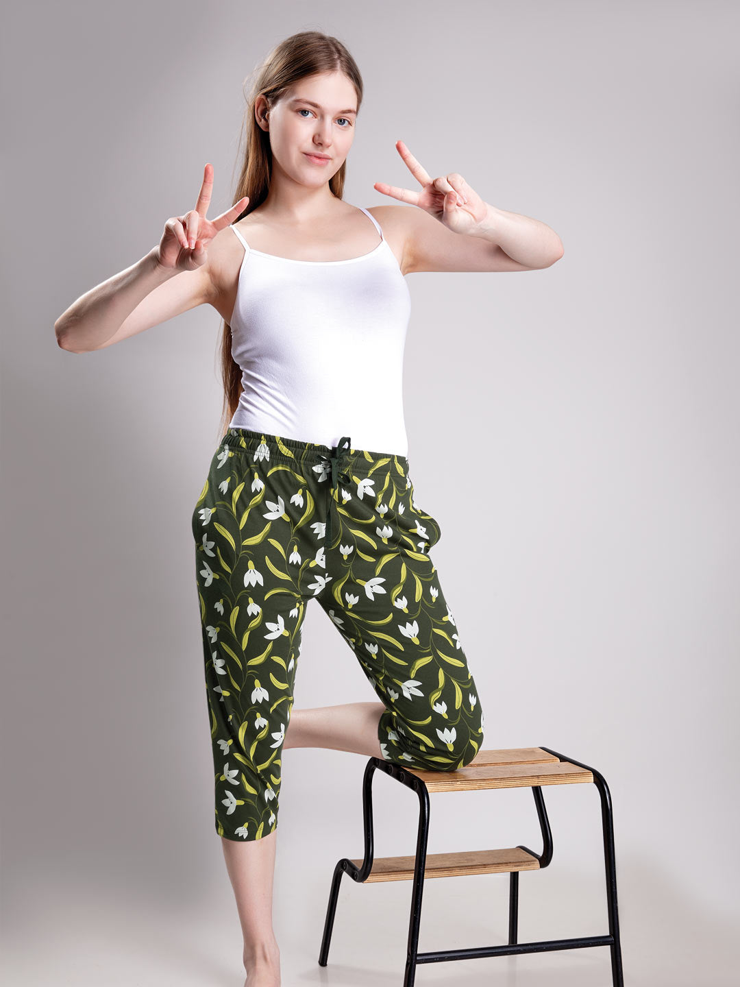 A woman posing in a white tank top and green capri pants with a white and yellow floral pattern, standing next to a small wooden step stool against a light gray background.