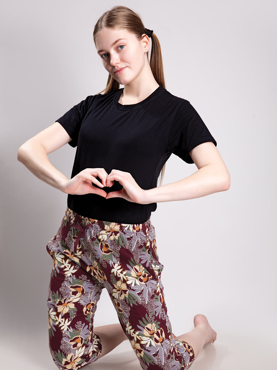 A woman kneeling and posing in a black short-sleeve t-shirt and floral print pants against a light gray studio background.