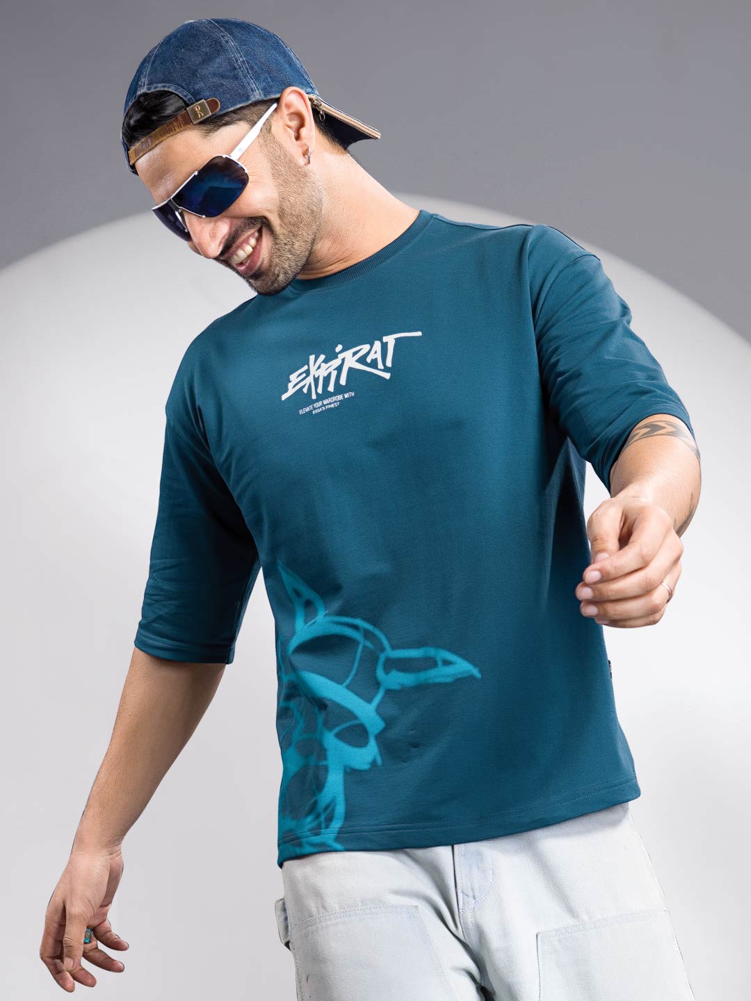 Man wearing a teal t-shirt with a logo and sunglasses, standing against a gray background