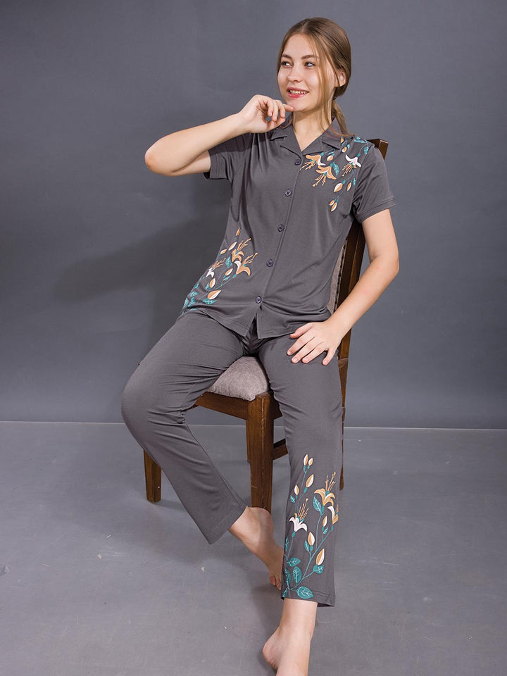 Woman wearing a gray pajama set with floral embroidery sitting on a chair against a gray background