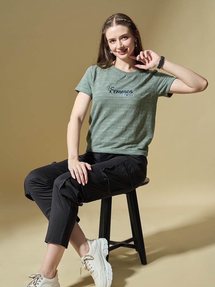 Woman sitting on a stool wearing a green t-shirt with a logo, black pants, and white sneakers on a beige background.