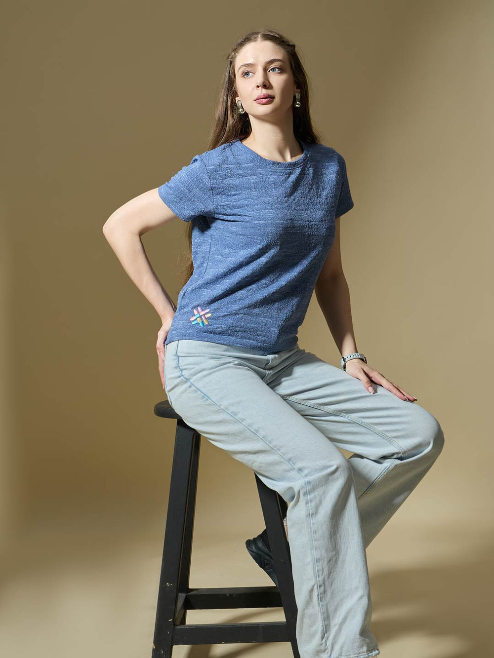 Woman sitting on a stool wearing a blue t-shirt and light blue jeans against a beige background