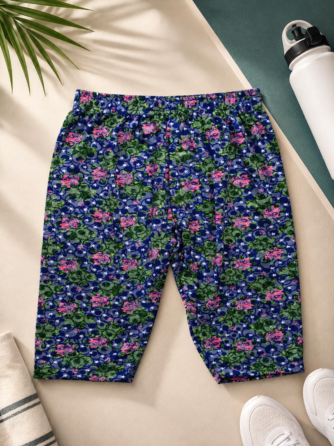 A pair of women's athletic shorts with a dense floral and paisley print in navy blue, green, and pink, laid flat on a beige surface with lifestyle props including a water bottle, sneakers, and a palm leaf.