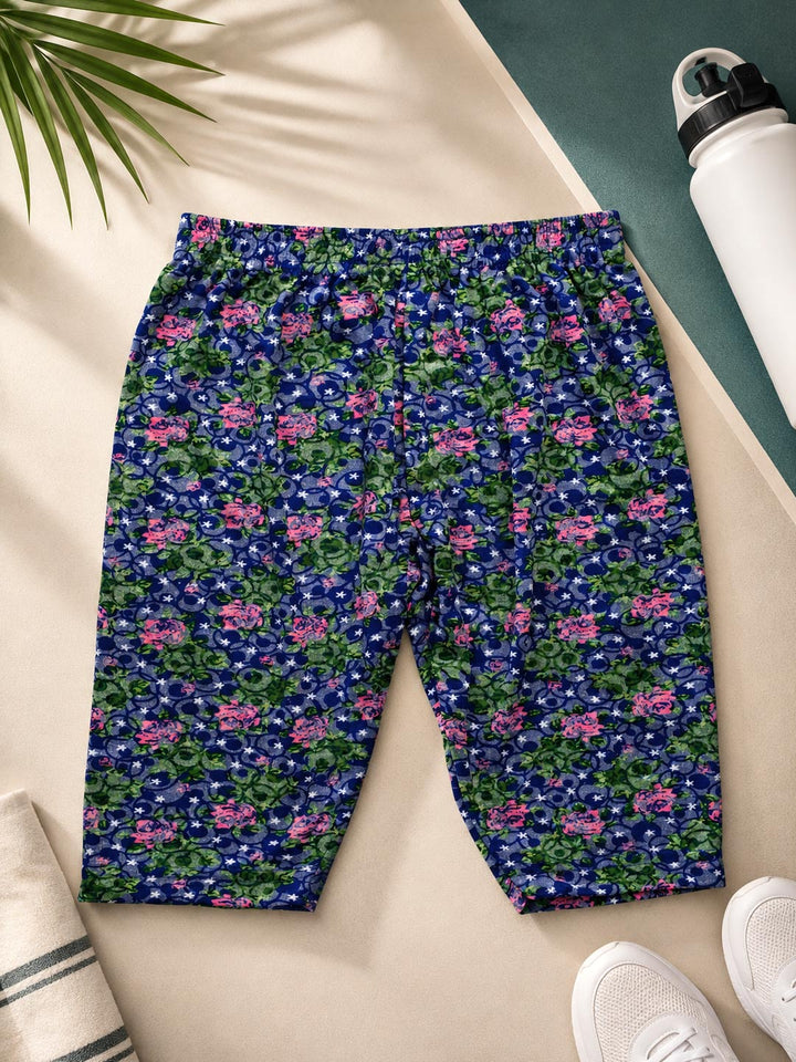 A pair of women's athletic shorts with a dense floral and paisley print in navy blue, green, and pink, laid flat on a beige surface with lifestyle props including a water bottle, sneakers, and a palm leaf.
