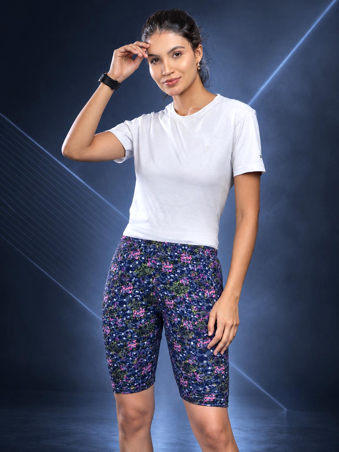 A woman modeling a white short-sleeve athletic top and navy blue biker shorts with a pink and green floral pattern, posed against a dark blue studio background with light streaks.
