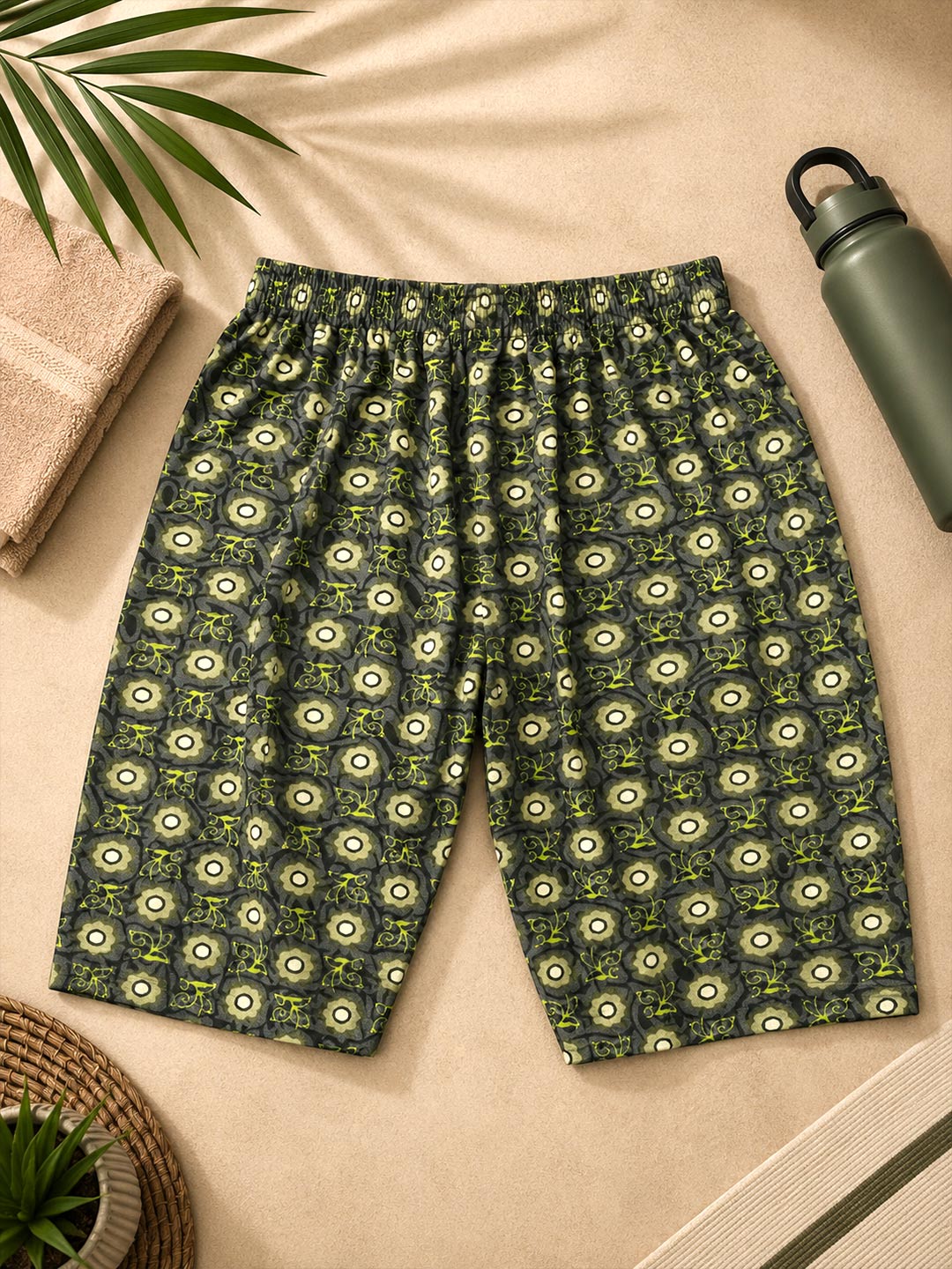A pair of casual shorts with an all-over circular eye-like pattern in olive green, cream, and dark gray, laid flat on a beige fabric background with a towel, plant leaf, and water bottle as props.