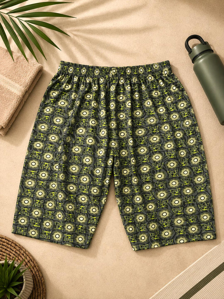 A pair of casual shorts with an all-over circular eye-like pattern in olive green, cream, and dark gray, laid flat on a beige fabric background with a towel, plant leaf, and water bottle as props.