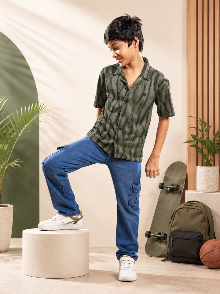 A boy modeling a short-sleeve button-up shirt with a green and black tie-dye pattern, paired with blue cargo pants and white sneakers, standing on a white cylindrical pedestal in a styled indoor setting with a skateboard, backpack, and potted plants. #color_rifle-green