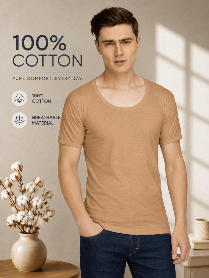 A man wearing a tan 100% cotton scoop neck t-shirt and dark blue jeans, standing in a warm, sunlit room with decorative cotton plants in a vase. Text overlays on the left highlight the product's cotton quality. calyx vest roundneck sleeve for men light brown-essa garments#color_harvest-gold
