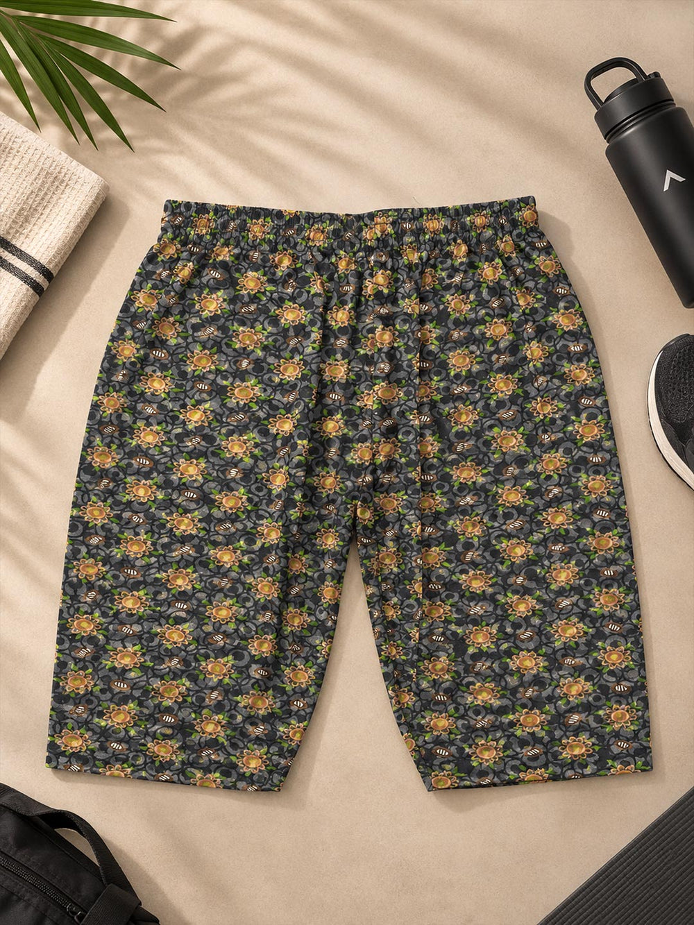 A pair of cropped shorts with an all-over small floral print in yellow, orange, and green on a dark navy background, laid flat on a beige surface with lifestyle props including a water bottle, a hat, and a shoe.