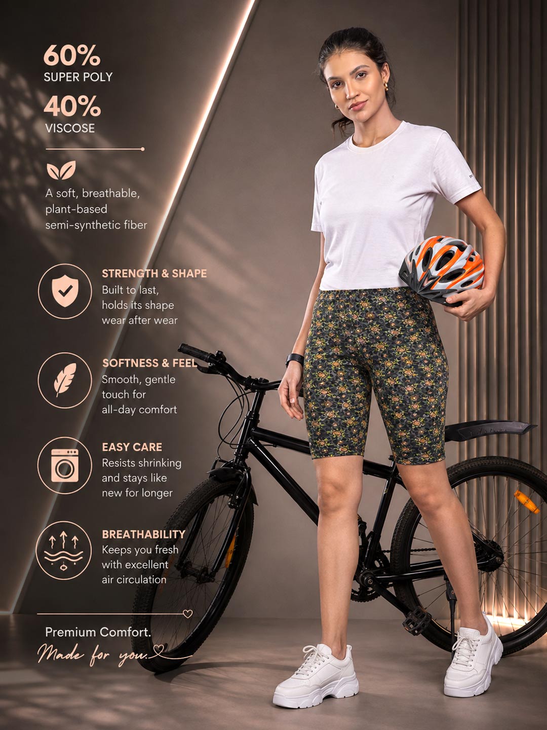 A woman stands beside a bicycle wearing patterned green bike shorts, a white T-shirt, and white sneakers, holding an orange and black bicycle helmet. Text on the left describes the shorts' material and benefits.
