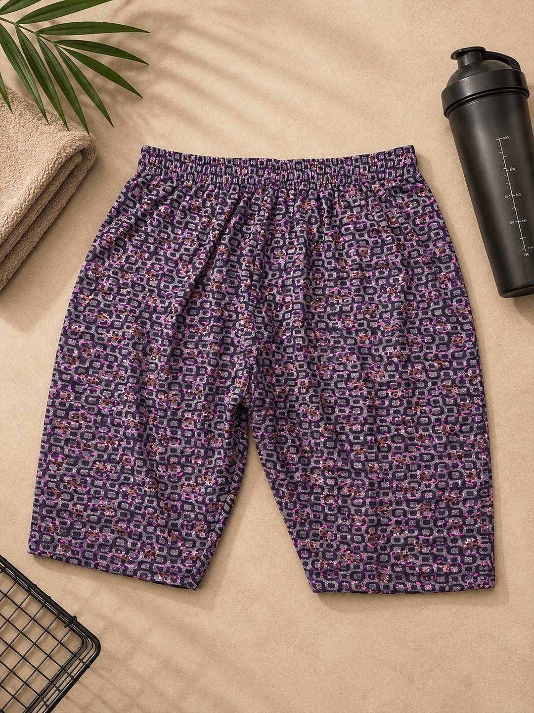 Purple and pink patterned athletic shorts laid flat on a beige surface with a black shaker bottle, folded towel, and plant leaves as props.