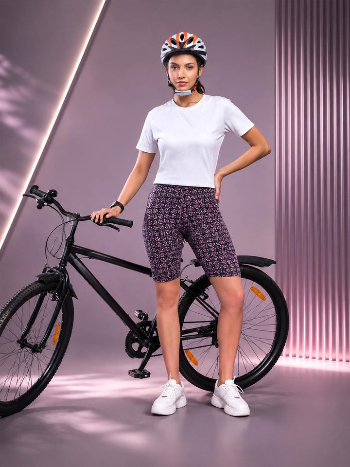 A woman wearing a white t-shirt, patterned purple and black biker shorts, white sneakers, and a bicycle helmet stands next to a black bicycle in a stylized studio setting with purple walls.