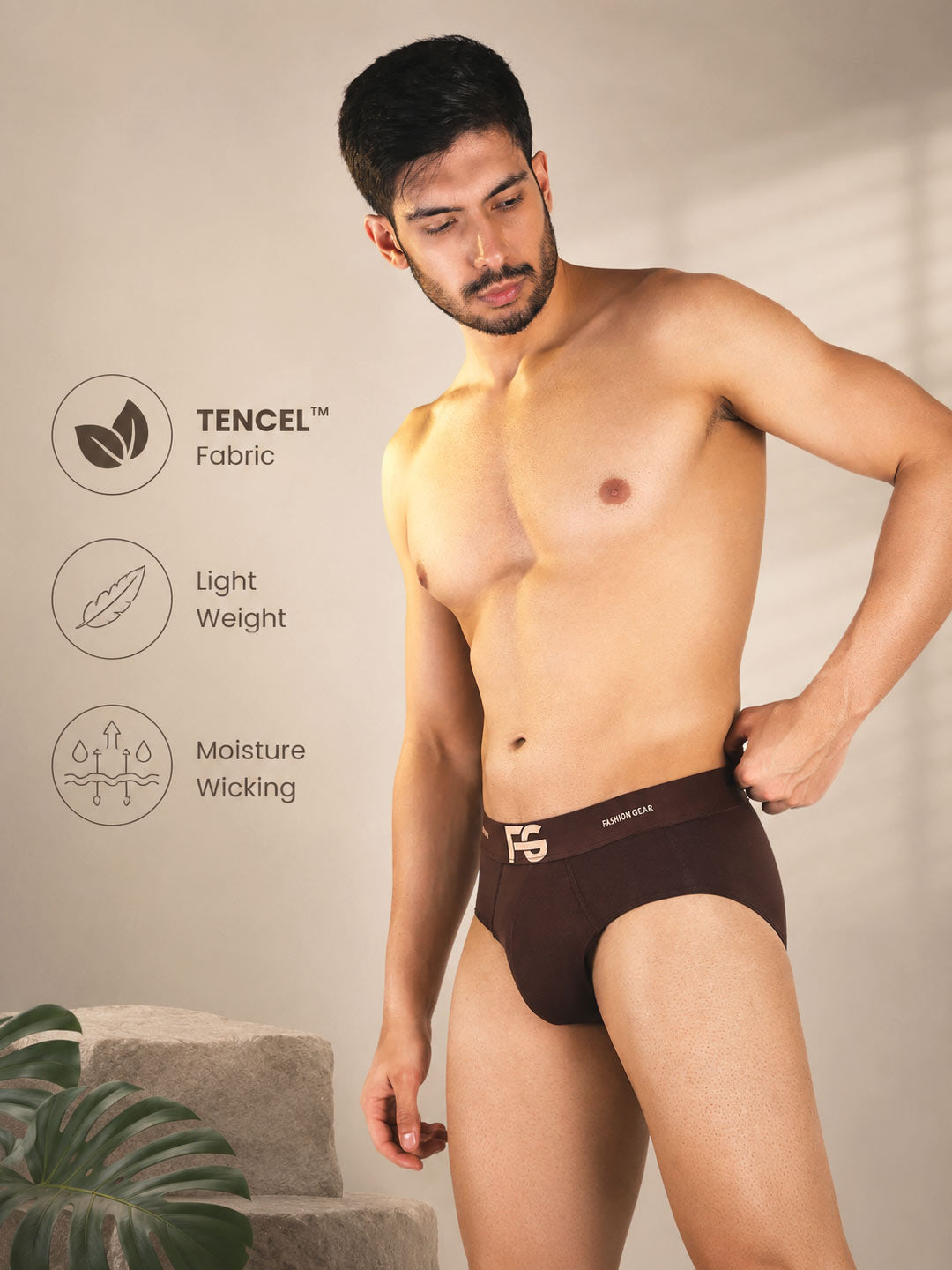 A shirtless man modeling dark brown briefs with a white logo, standing against a warm beige wall with product feature icons and text on the left side.#color_irish-coffee