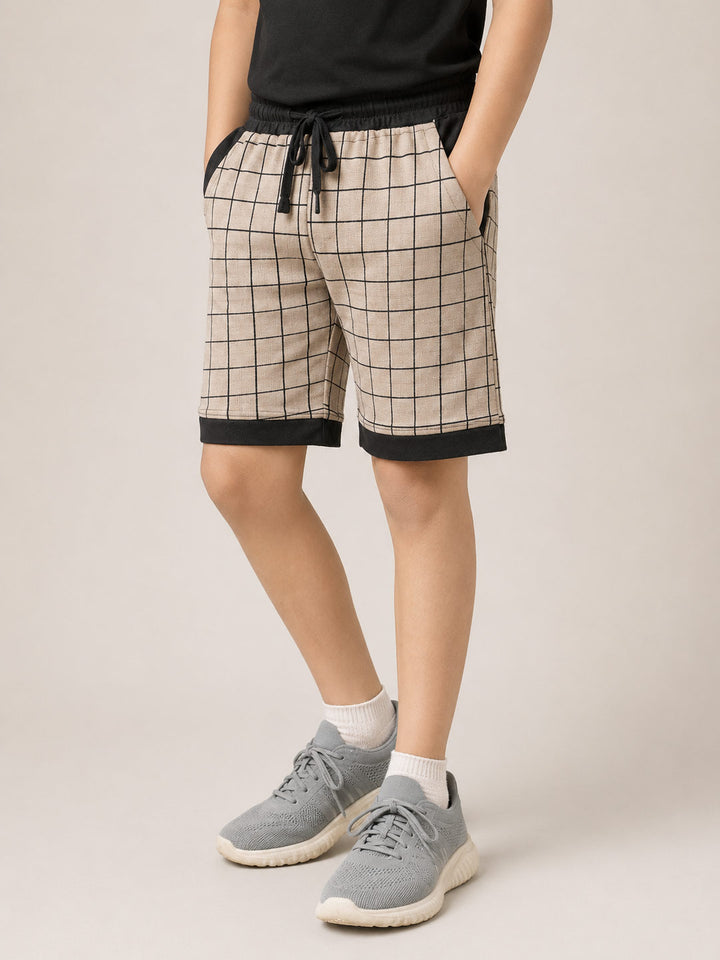 A person wearing beige plaid shorts with black trim and a black drawstring, paired with a black top, white socks, and gray sneakers, standing against a light beige studio background.fitzone-105 shorts for boys color_black
