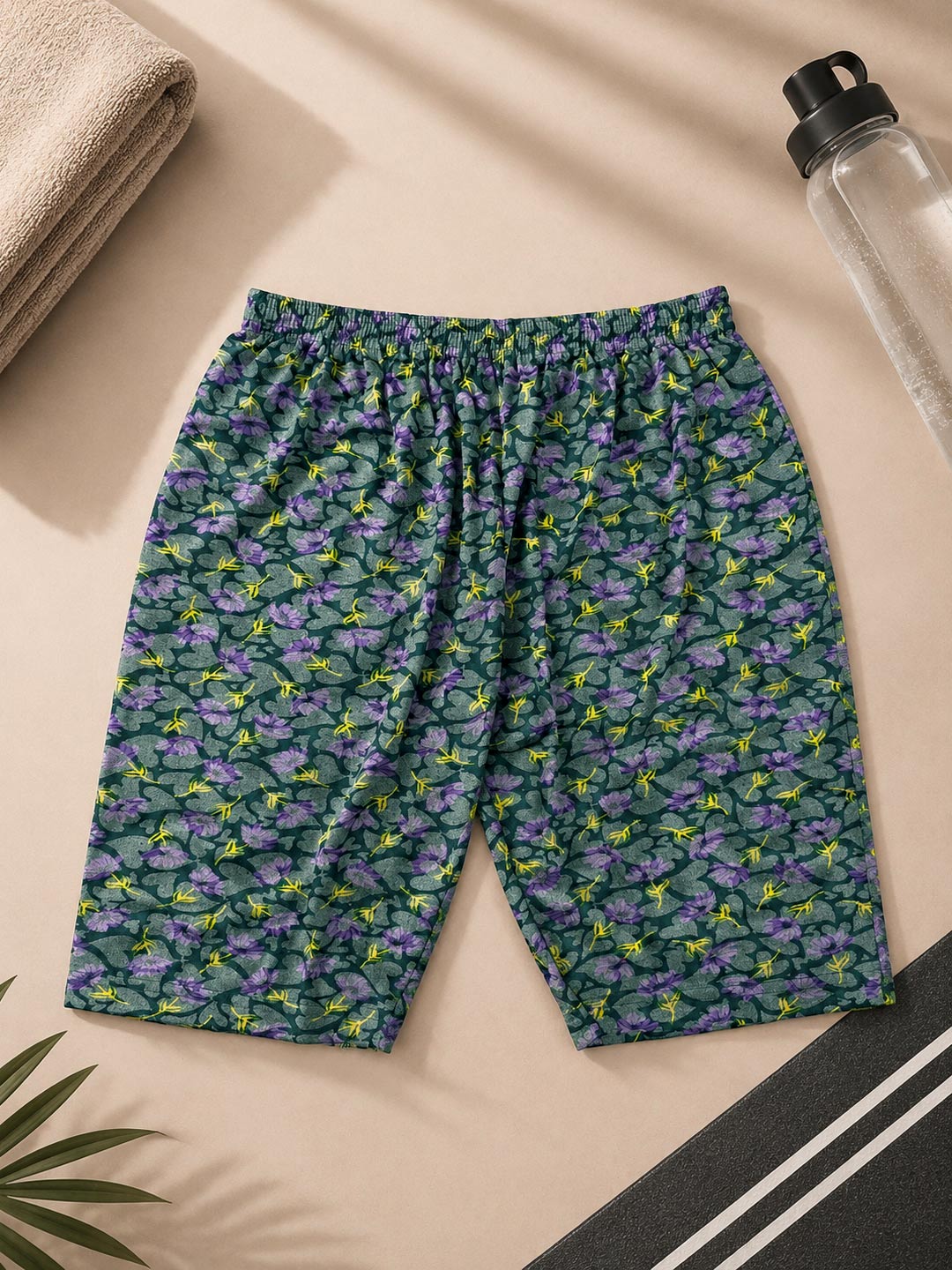 A pair of athletic shorts with an all-over butterfly print in purple, green, and yellow, laid flat on a beige background with lifestyle props including a towel, a water bottle, and a plant leaf.
