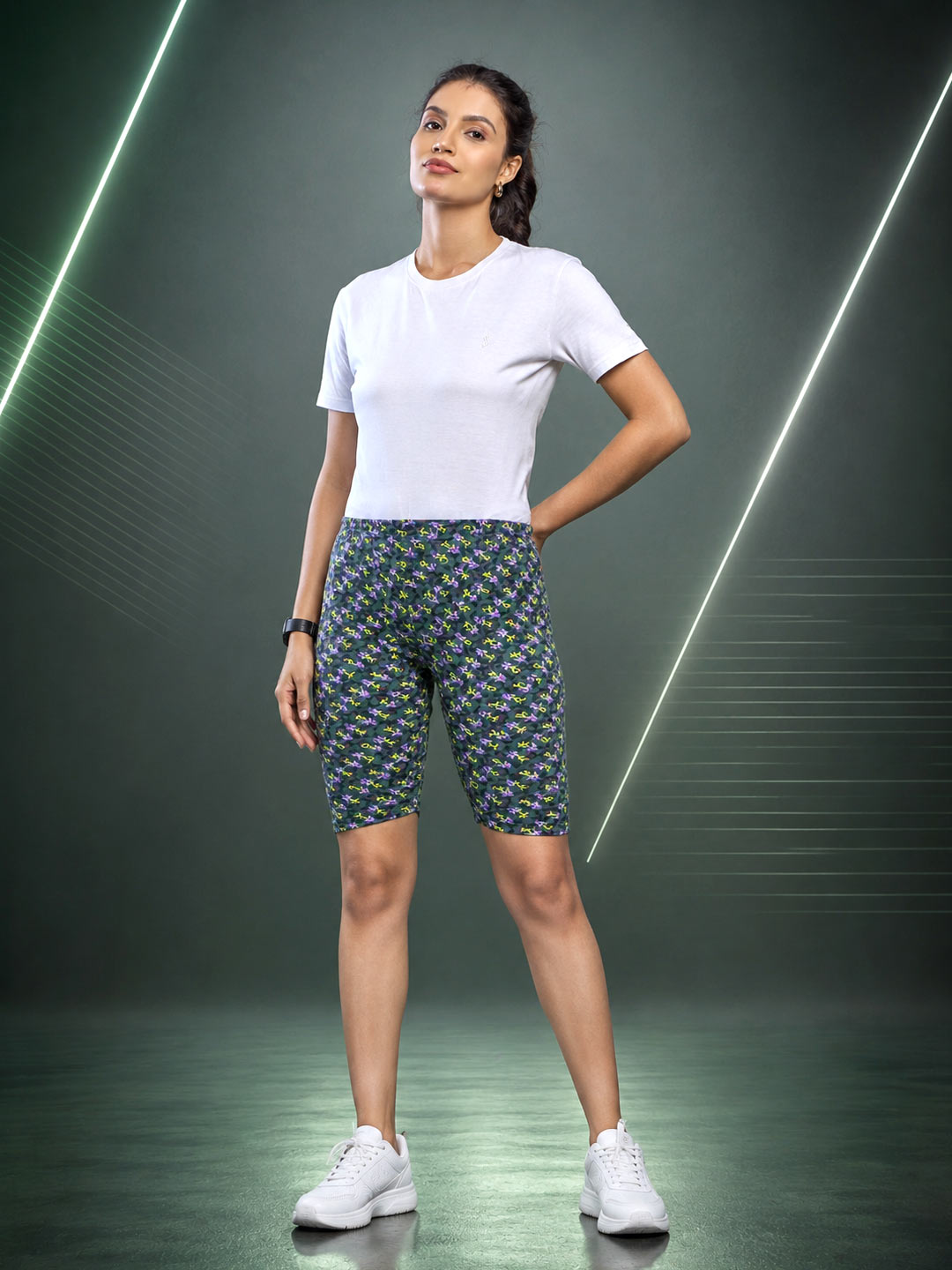 A woman modeling fitted biker shorts with a dense small-scale floral pattern in green, yellow, and blue, paired with a plain white t-shirt and white sneakers, set against a dark studio background with glowing diagonal light lines.