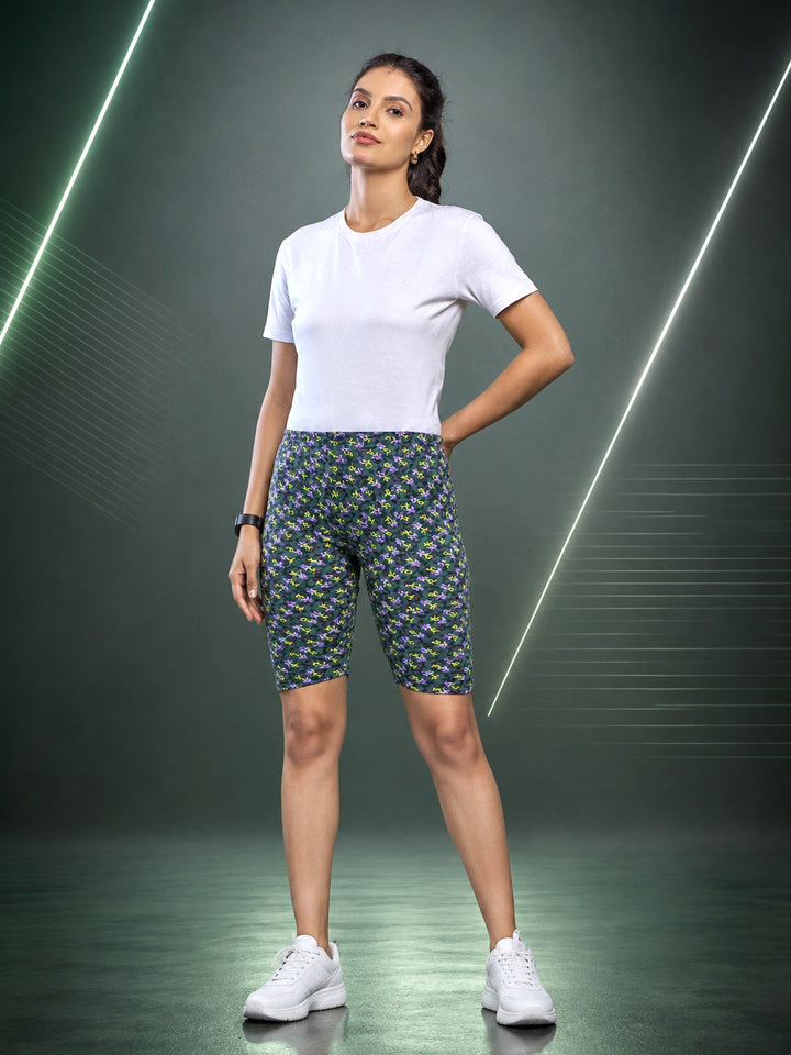 A woman modeling fitted biker shorts with a dense small-scale floral pattern in green, yellow, and blue, paired with a plain white t-shirt and white sneakers, set against a dark studio background with glowing diagonal light lines.