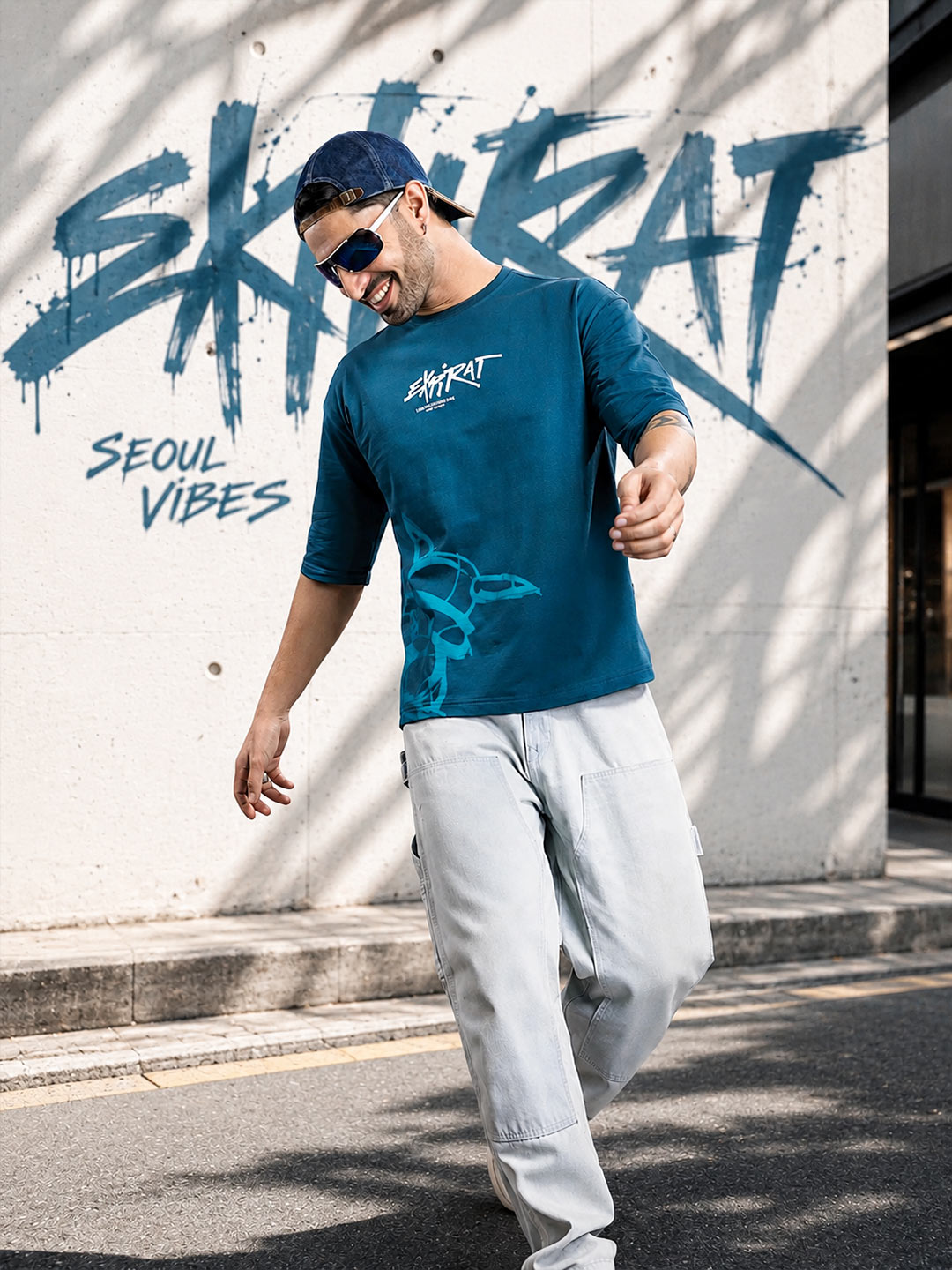 A man wearing a teal blue 3/4 sleeve graphic t-shirt with 'SKYRAT' and 'SEOUL VIBES' text, paired with light gray cargo pants and a navy cap, walking outdoors against a concrete wall with graffiti.
