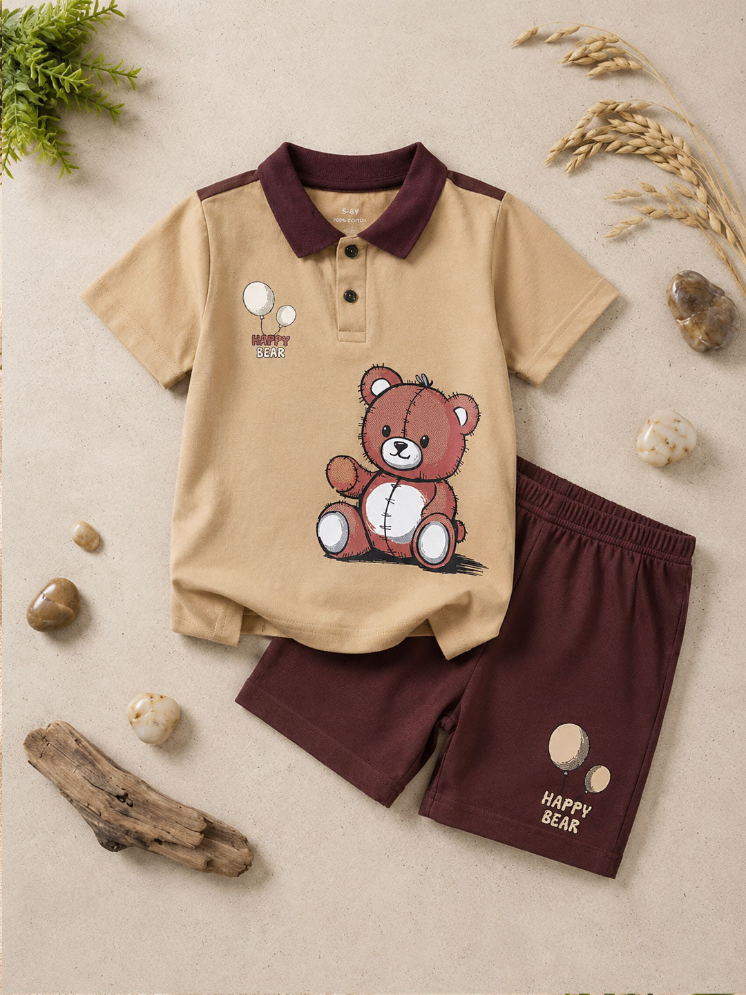 A children's two-piece clothing set laid flat on a beige textured surface with decorative stones, driftwood, and dried grass. The set includes a short-sleeve polo shirt in beige with a dark burgundy collar and a brown teddy bear graphic on the front, paired with matching dark burgundy shorts featuring a small teddy bear graphic and 'HAPPY BEAR' text.