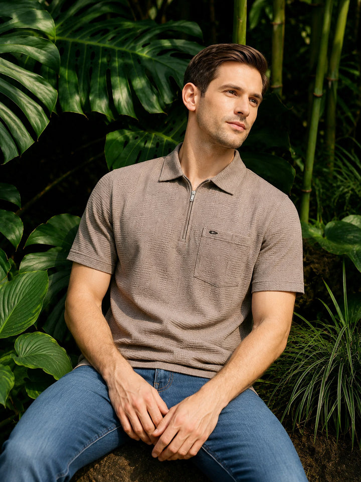 A man wearing a taupe brown short-sleeve polo shirt with a half-zip collar and blue jeans, sitting in front of a lush background of green tropical leaves and bamboo.