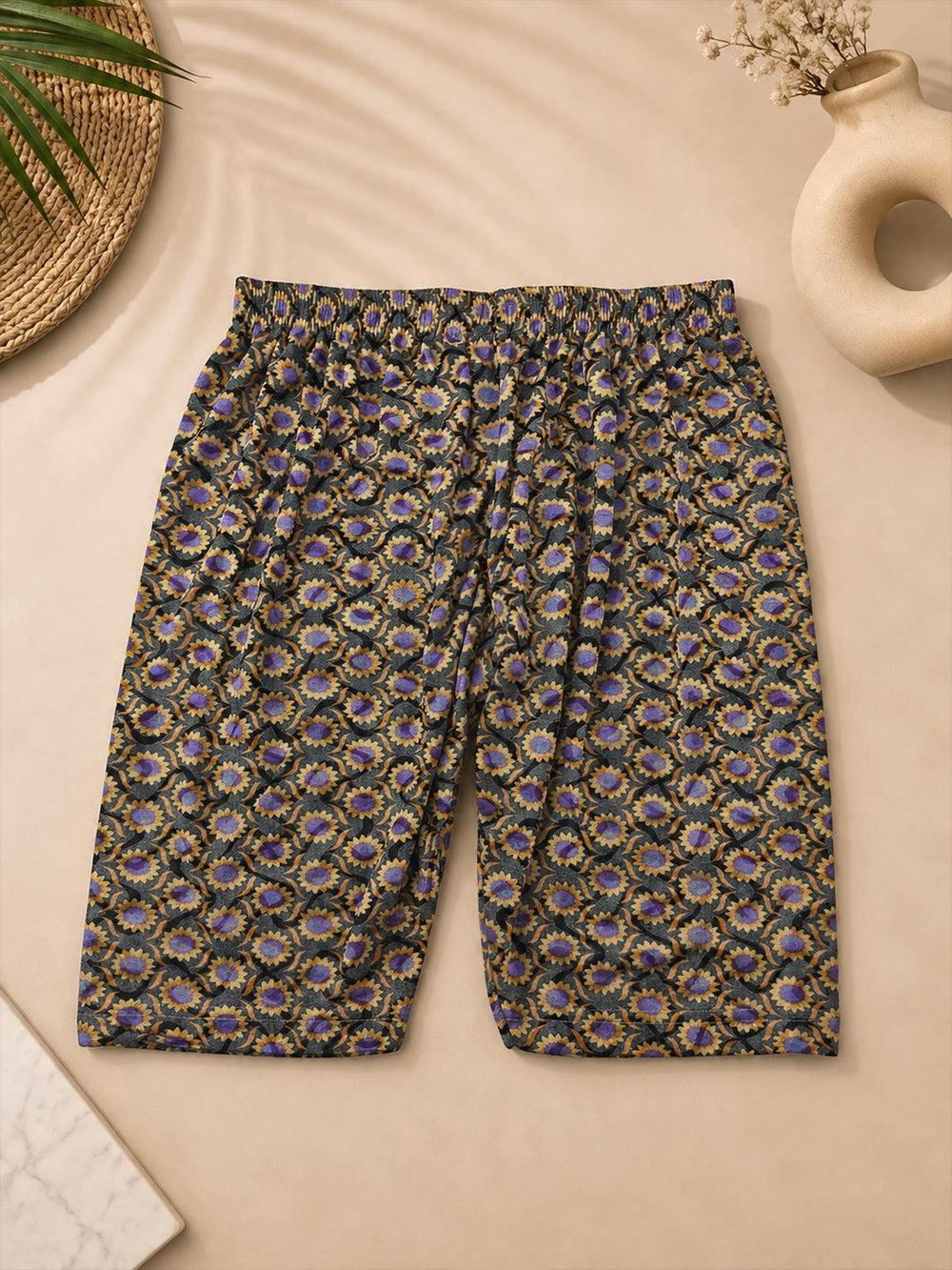 A pair of women's cropped shorts with an elastic waistband, featuring an all-over intricate paisley and floral pattern in purple, gold, and green on a black base, laid flat on a beige background with styled props.