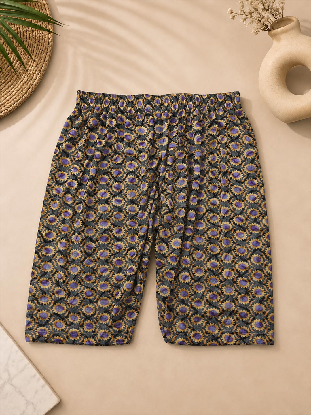 A pair of women's cropped shorts with an elastic waistband, featuring an all-over intricate paisley and floral pattern in purple, gold, and green on a black base, laid flat on a beige background with styled props.