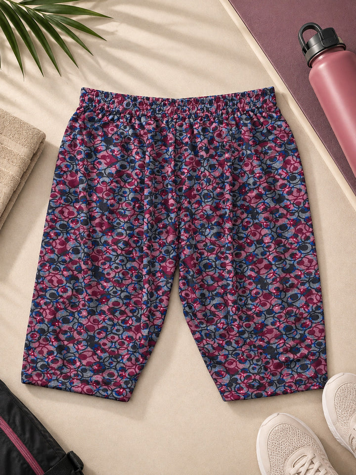 A pair of women's cropped leggings with an all-over abstract floral pattern in pink, purple, and blue, laid flat on a beige surface with lifestyle props including a pink water bottle, white sneakers, and a plant leaf.