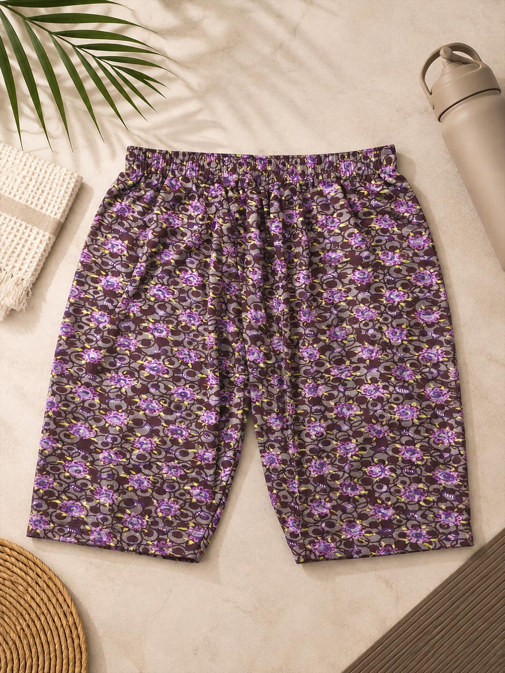 A pair of women's shorts with an all-over purple and black floral pattern, laid flat on a light beige textured surface with decorative props including a palm leaf, a woven towel, a straw hat, and a water bottle.