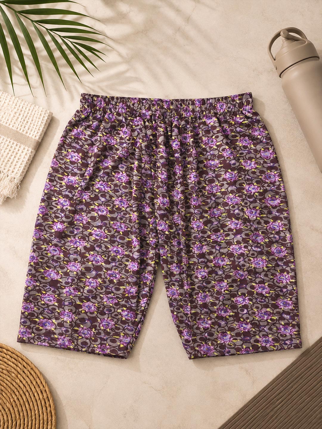 A pair of women's shorts with an all-over purple and black floral pattern, laid flat on a light beige textured surface with decorative props including a palm leaf, a woven towel, a straw hat, and a water bottle.