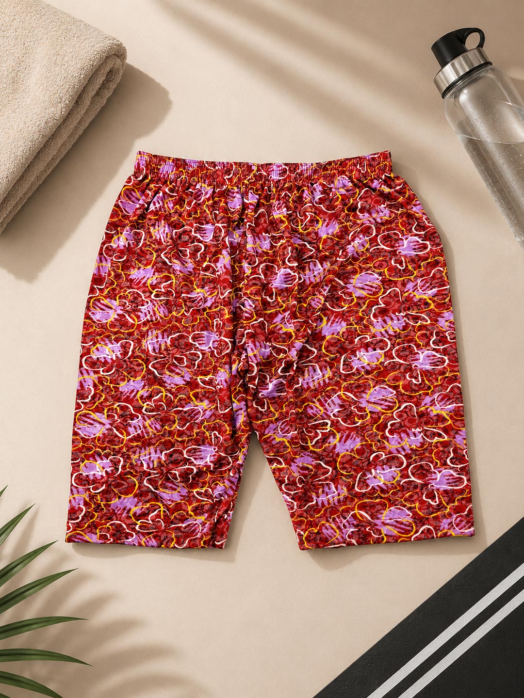 Red and pink patterned swim trunks laid flat on a beige surface with a towel, water bottle, and plant leaves as props.