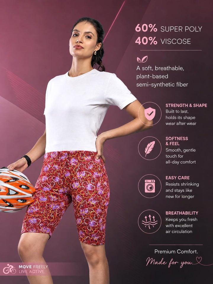 A woman modeling patterned athletic shorts and a white T-shirt on a dark pink promotional background with text describing the fabric and product benefits. The shorts have a red base with a dense floral and paisley print in pink, purple, and orange.