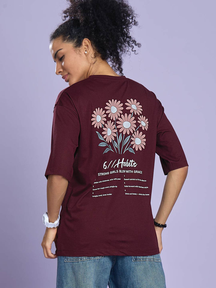 Person wearing a maroon t-shirt with floral design and text on the back, standing against a light purple background.