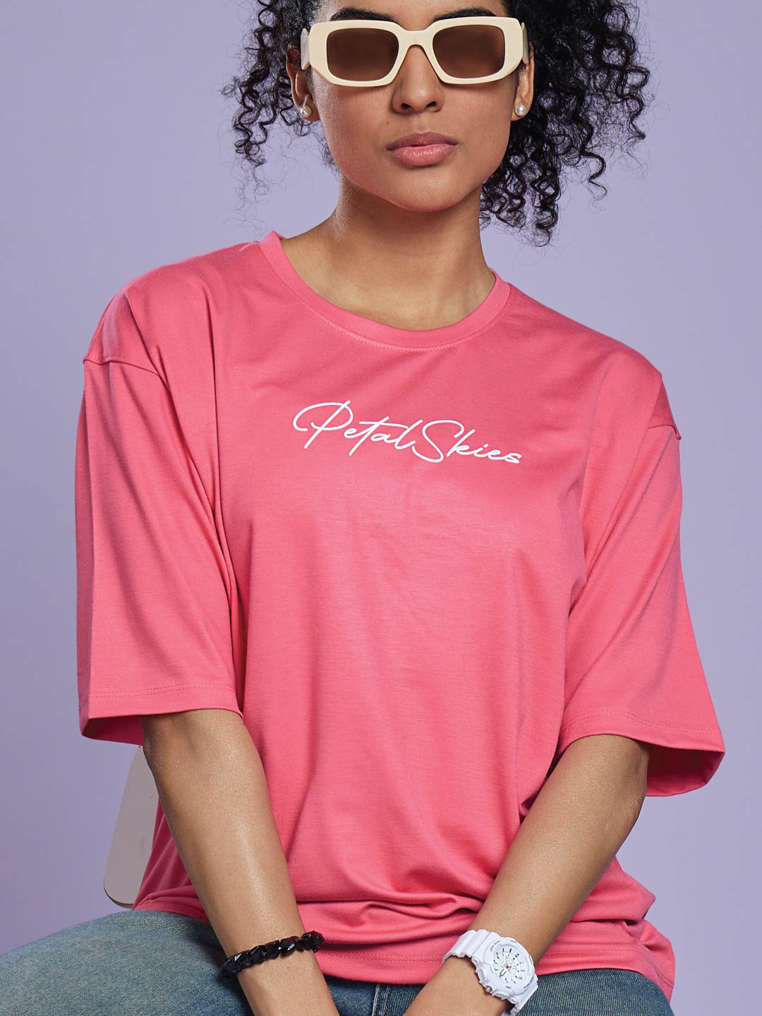 Person wearing a pink t-shirt with 'Petals & Shinies' text, sunglasses, and a watch on a purple background