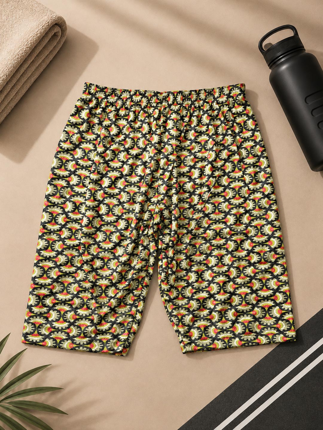 A pair of women's athletic shorts with an all-over abstract floral pattern in orange, yellow, white, and green on a black background, laid flat on a beige surface with lifestyle props including a water bottle, towel, and plant.